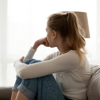 Side view young woman looking away at window sitting on couch at home. Frustrated confused female feels unhappy