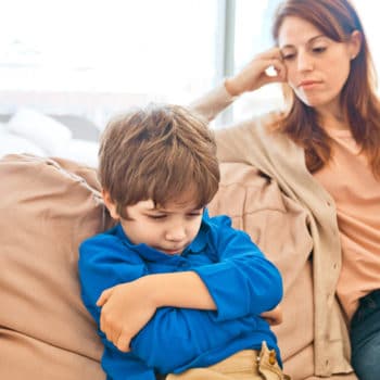 Displeased son with anger issues sitting with arms crossed on sofa at home. Worried mother looking at her child. Focus on boy.