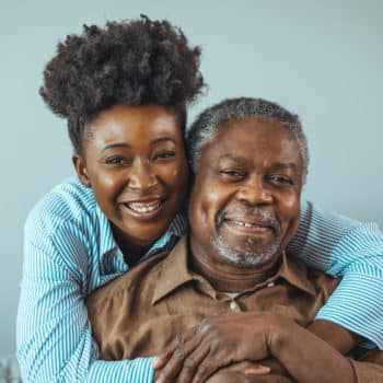 Senior man and his middle aged daughter smiling at each other embracing, close up. Portrait of a daughter holding her elderly father, sitting on a bed by a window in her father's room.